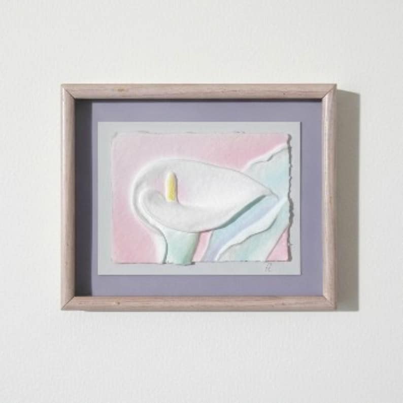 May include: A framed artwork featuring a textured, embossed paper relief of a white calla lily with a yellow spadix. The flower is set against a soft pink and light blue background with deckled edges, enclosed in a light wood frame with a lavender mat.