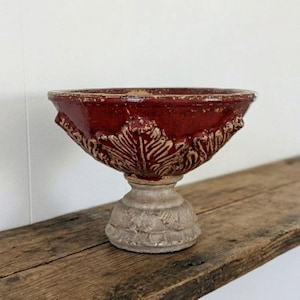 May include: A decorative, antique-style urn with a deep red, textured bowl and a light beige pedestal base. The bowl features embossed leaf designs and is set on a weathered wooden surface.