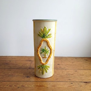 May include: A tall, off-white ceramic vase with a tropical theme. The vase features a central diamond-shaped design with a palm tree and bamboo-style trim. Green and yellow accents highlight the palm leaves and frame. The vase stands on a wooden surface.