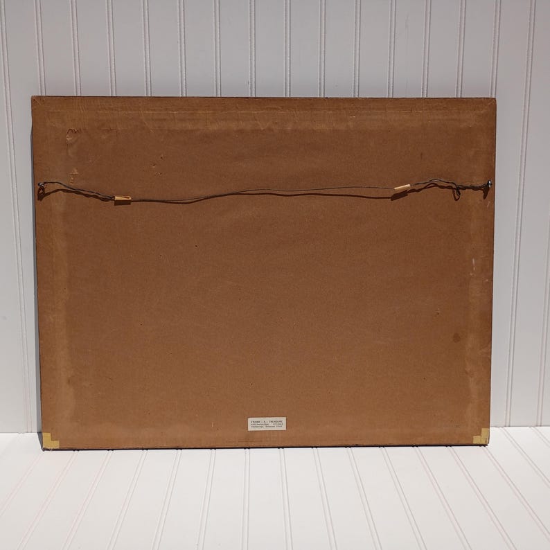 Op de afbeelding: Achterkant van een ingelijst kunstwerk. De lijst is donkerbruin en de achterkant is lichtbruin karton. Een draad is over de achterkant gespannen om op te hangen. Goudkleurige hoekbeschermers zijn zichtbaar.