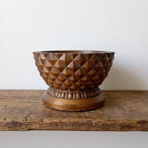 May include: A brown wooden bowl with a geometric diamond pattern. The bowl sits on a pedestal base with a decorative ring of small vertical lines. The bowl is displayed on a rustic wooden shelf.