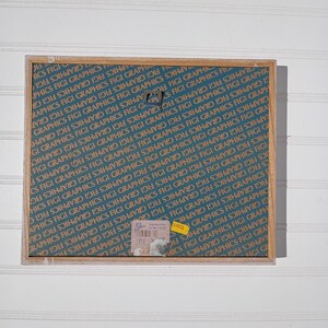 May include: A rectangular wooden frame with a teal background featuring repeating text that reads "GRAPHICS FIGI". The frame has a metal hook and several stickers attached. The overall aesthetic is vintage.