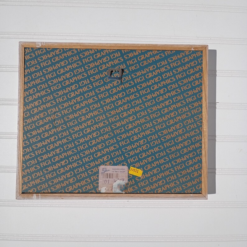 May include: A rectangular wooden frame with a teal background featuring repeating text that reads "GRAPHICS FIGI". The frame has a metal hook and several stickers attached. The overall aesthetic is vintage.
