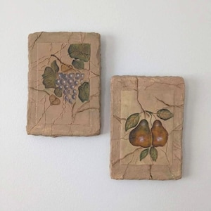 May include: Two rectangular wall art pieces with a textured, cracked appearance. One features a bunch of purple grapes and green leaves, the other depicts two brown pears with green leaves. The artwork has a neutral color palette.