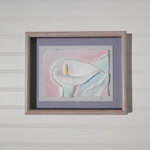 May include: Framed artwork featuring a white calla lily with a yellow stamen, set against a pink and blue watercolor background. The artwork is in a light-colored wooden frame, creating a soft, artistic aesthetic.