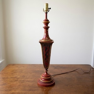 May include: A vintage table lamp with a red base and body, accented with gold trim and black floral designs. The lamp has a brass bulb socket and a brown electrical cord. The lamp stands approximately 28 inches tall.