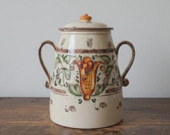 May include: Cream-colored ceramic jar with lid and two handles. The jar features a floral design in orange, green, and brown. The lid has a small orange knob. The jar is on a wooden surface.