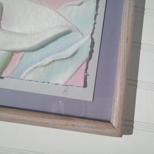 May include: Abstract watercolor painting in a light wood frame. The artwork features soft pastel colors, including white, pink, blue, and green. The painting has a textured appearance and is signed in the lower right corner. The frame has a light, natural wood finish.