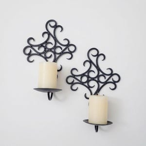 May include: A pair of black metal wall sconces featuring intricate scrollwork designs. Each sconce includes a circular platform holding a cylindrical off-white pillar candle. The metal frames have a symmetrical, openwork pattern with curved lines.