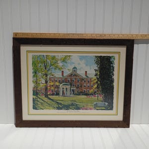 Op de afbeelding: Ingelijste prent van een universiteitsgebouw met een koepeldak en een witte portiek. De prent zit in een donkerbruine houten lijst. De afbeelding bevat bomen, een bank en de tekst "The Old Well and South Building. The University of North Carolina at Chapel Hill."