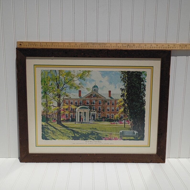 Op de afbeelding: Ingelijste prent van een universiteitsgebouw met een koepeldak en een witte portiek. De prent zit in een donkerbruine houten lijst. De afbeelding bevat bomen, een bank en de tekst "The Old Well and South Building. The University of North Carolina at Chapel Hill."