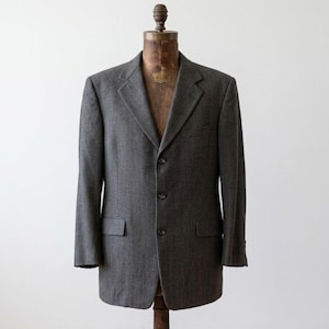 May include: A charcoal gray blazer with a three-button closure, displayed on a wooden dress form. The blazer has two front pockets and a classic notched lapel. The fabric appears to be a textured wool blend, suitable for a professional or formal setting.