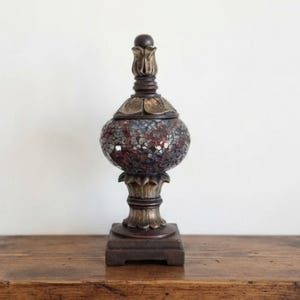 May include: A decorative urn featuring a central bulbous body covered in a mosaic of dark red, grey, and black glass tiles. The base and the removable lid are crafted from a dark brown material with a metallic gold finish on the edges.