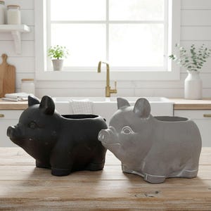 May include: Two ceramic pig planters, one black and one gray, sit side-by-side on a wooden surface. The planters have open tops for plants and are designed with pig features, including ears, snouts, and small legs. The background shows a kitchen setting.