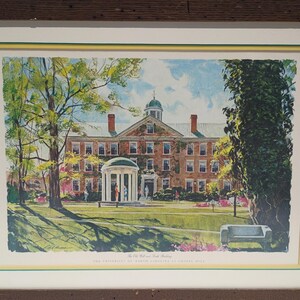 Op de afbeelding: Een ingelijste prent van de Old Well en het hoofdgebouw van de Universiteit van North Carolina in Chapel Hill. De prent toont een gedetailleerde aquarel van de campus met bomen, een prieel en een bakstenen gebouw. De prent is ingelijst in een bruine houten lijst.