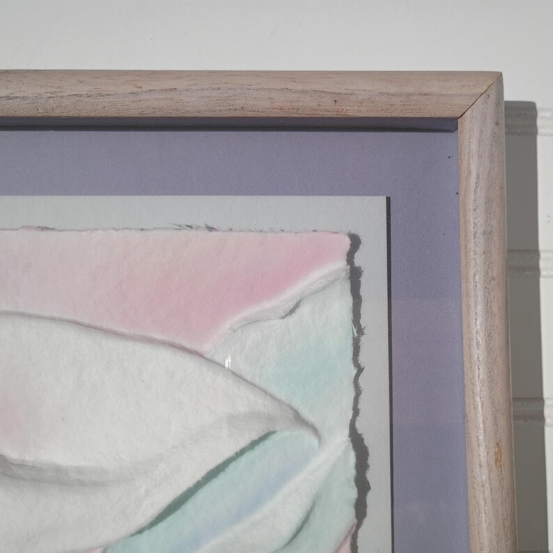 May include: A framed artwork featuring textured, layered white paper with soft pink and light blue watercolor accents. The piece is set against a lavender mat board and enclosed in a light-toned wooden frame with a visible grain pattern.