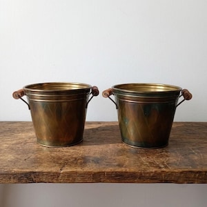 May include: A pair of matching brass buckets featuring a tapered cylindrical shape with a subtle diamond-patterned texture on the exterior. Each bucket is equipped with two metal side handles that are accented with small, cylindrical wooden grips.