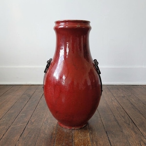 May include: A tall, red glazed ceramic vase with a wide body and a slightly flared rim. The vase has two dark metal handles and sits on a wooden floor. The vase is approximately 24 inches tall.