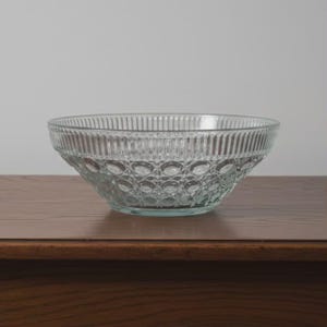 May include: A clear glass bowl with a textured design. The bowl has a ribbed pattern around the top and a repeating oval pattern on the sides. The bowl is sitting on a wooden surface.