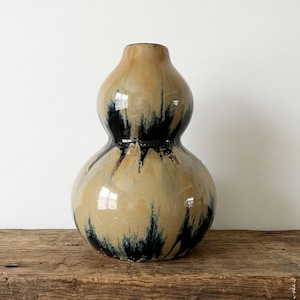 May include: A gourd-shaped ceramic vase featuring a glossy finish with a tan base color and dark blue, drip-like glaze patterns that cascade from the neck down to the rounded body, creating a distinct contrast across the smooth surface.