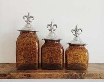 May include: Three amber-colored glass canisters of varying heights, each featuring a square base and a removable silver-toned metal lid topped with a decorative fleur-de-lis finial. The glass has a mottled, tortoiseshell-like pattern throughout.
