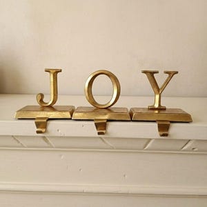 May include: Gold-colored metal stocking holders spelling out the word "JOY". Each letter is attached to a rectangular base with a hook for hanging stockings. The letters and bases have a metallic finish, creating a festive holiday decoration.