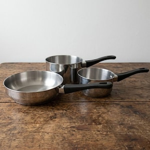 May include: Three stainless steel saucepans with black handles are arranged on a rustic wooden surface. The pans vary in size, with a small frying pan and two saucepans of different heights. The shiny metal reflects the light, creating a clean, modern look.