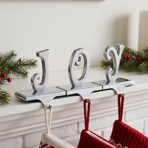 May include: A silver-toned metal stocking holder set spelling the word JOY. Each letter is mounted on a rectangular base with a hook underneath. The letters have a polished finish and are designed to sit on a mantel edge to hold hanging stockings.