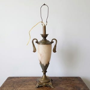 May include: A table lamp featuring a central urn-shaped body made of smooth, cream-colored stone. The lamp has ornate brass-toned metal handles, a decorative base with leaf-like detailing, and a metal harp fixture extending from the top.