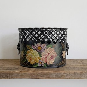 May include: A black cylindrical metal container featuring a decorative lattice pattern along the top rim and hand-painted floral motifs in shades of yellow, pink, and green. The vessel includes two small metal ring handles on the sides.