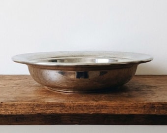 May include: A shallow, circular silver-toned metal bowl with a wide, flat rim. The bowl features a polished, reflective surface with subtle signs of wear and a smooth, rounded base, presenting a simple, functional design for serving or display.