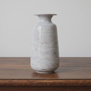 May include: A white and gray marbled ceramic vase with a flared top and a rounded base. The vase has a cylindrical shape and is decorated with streaks of red and blue. The vase is sitting on a wooden surface.