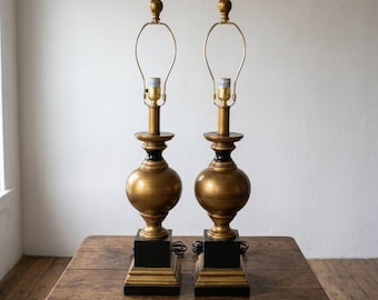 May include: Two matching gold-toned table lamps with black accents and square bases. The lamps have a classic, elegant design with a bulbous body and a brass-colored neck and finial. They sit on a wooden table.