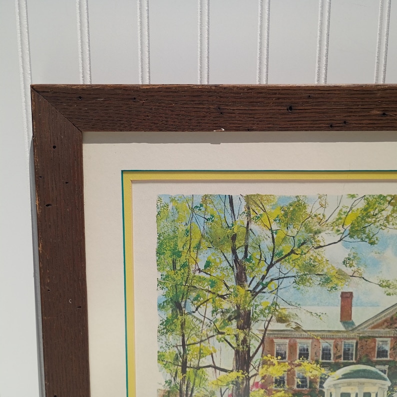 Op de afbeelding: Een ingelijste prent met een aquarel van een gebouw met een rood bakstenen dak en een witte koepel. Het kunstwerk is omgeven door groene bomen en een gele en groene rand, in een donkerbruine houten lijst.