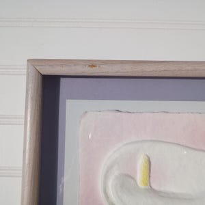 May include: A framed artwork featuring a white, abstract form with a yellow accent on a pink background. The artwork is set within a light gray mat and a light wood frame. The piece is mounted on a white wall.