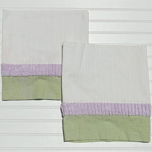May include: Two white pillowcases with vertical stripes. Each pillowcase has a lavender ruffled trim and a green gingham patterned bottom border. The pillowcases are displayed on a white, horizontal-slatted surface.