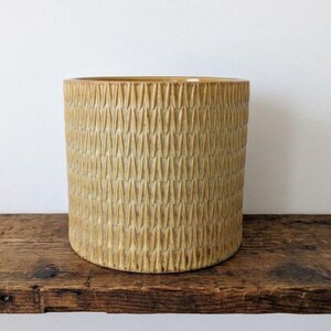 May include: A cylindrical ceramic planter featuring a mustard yellow glaze and a repeating vertical textured pattern of raised, leaf-like shapes covering the entire exterior surface of the vessel from the top rim to the base.