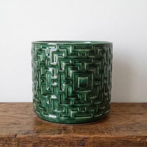 May include: A cylindrical ceramic planter featuring a deep forest green glaze and a raised, intricate geometric maze pattern that wraps around the entire exterior surface, creating a textured relief effect with glossy highlights.