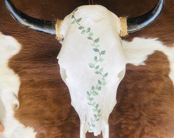 May include: White cow skull with black horns and a painted green vine design. The skull is mounted on a brown and white cowhide rug.