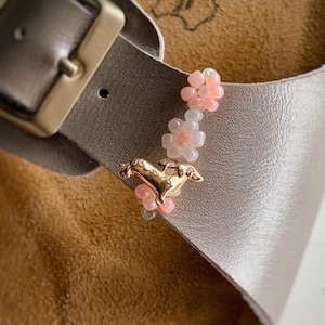 Könnte beinhalten: Ein dekorativer Anhänger an einem metallischen Sandalenriemen, bestehend aus einer goldfarbenen Dackel-Figur, die von kleinen, durchscheinenden rosa und weißen blütenförmigen Perlen flankiert wird, welche vertikal neben der Schnalle angeordnet sind.