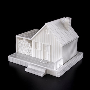 May include: A white 3D printed model of a small house with a wood-like exterior, a chimney, a door, and a window. The house has a small porch with steps and a woodpile on the side.