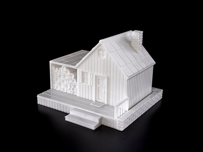 May include: A white 3D printed model of a small house with a wood-like exterior, a chimney, a door, and a window. The house has a small porch with steps and a woodpile on the side.