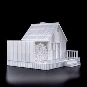May include: A white 3D printed model of a small house with a wood-like texture. The house has a roof, a door, a window, and a small porch. There is a stack of logs next to the house.