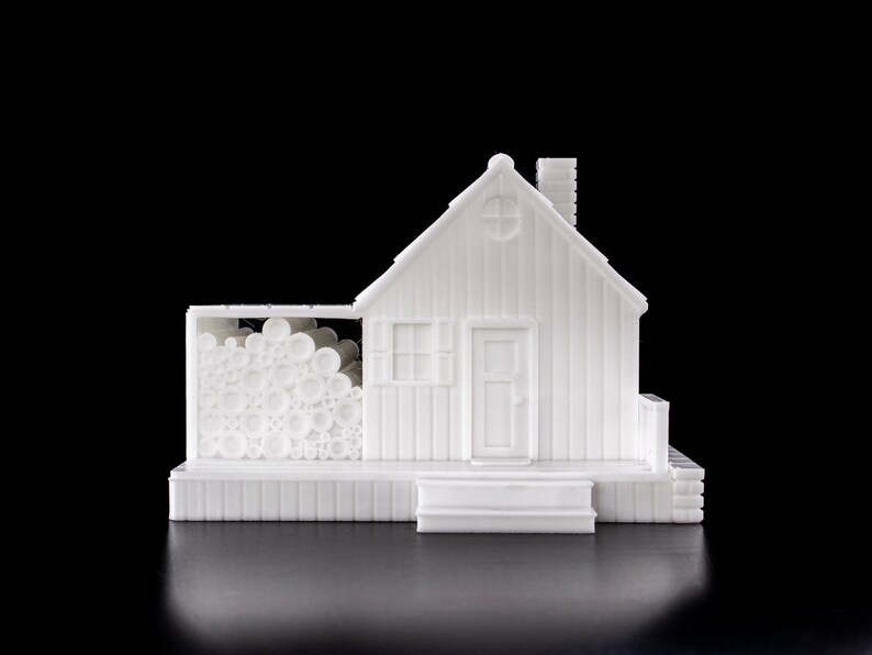 May include: White 3D printed model of a small house with a woodpile on the side. The house has a door, a window, and a chimney. The woodpile is stacked high and is made of small, round logs.