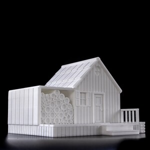 May include: A white 3D printed model of a small house with a wood-like texture. The house has a roof, a door, a window, and a porch. There is a pile of logs next to the house.