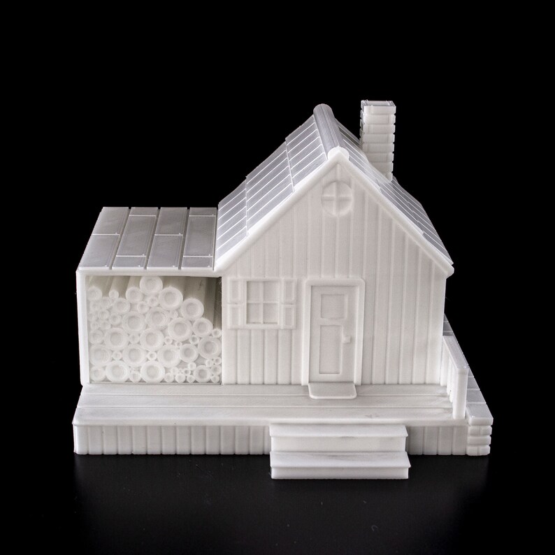 May include: A white 3D printed model of a small house with a wood-like texture. The house has a chimney, a door, and a window. There is a pile of logs in front of the house.