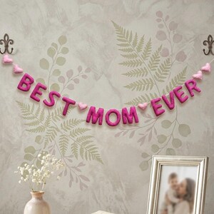 May include: A felt banner with the words "BEST MOM EVER" in magenta letters, strung with pink felt hearts. The banner hangs against a floral wallpaper background, with a framed photo and a vase of flowers below.