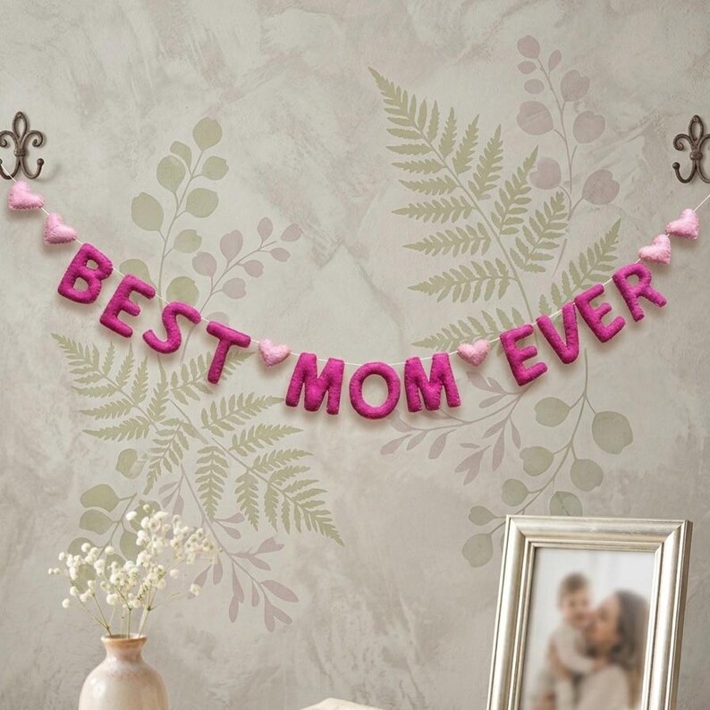 May include: A felt banner with the words "BEST MOM EVER" in magenta letters, strung with pink felt hearts. The banner hangs against a floral wallpaper background, with a framed photo and a vase of flowers below.