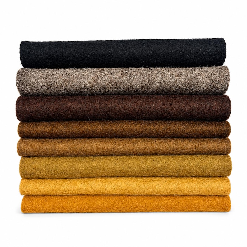 May include: A stack of eight rectangular felt sheets arranged in a gradient from black at the top, transitioning through shades of grey, dark brown, medium brown, light brown, ochre, and golden yellow at the bottom, featuring a dense, fibrous texture.
