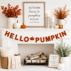Op de afbeelding: Een viltbanner met oranje letters die HELLO PUMPKIN spellen, inclusief een klein pompoenicoon in het midden. De display bevat witte en oranje pompoenen, herfsttakken in witte vazen, kaarsen en twee ingelijste borden met tekst.